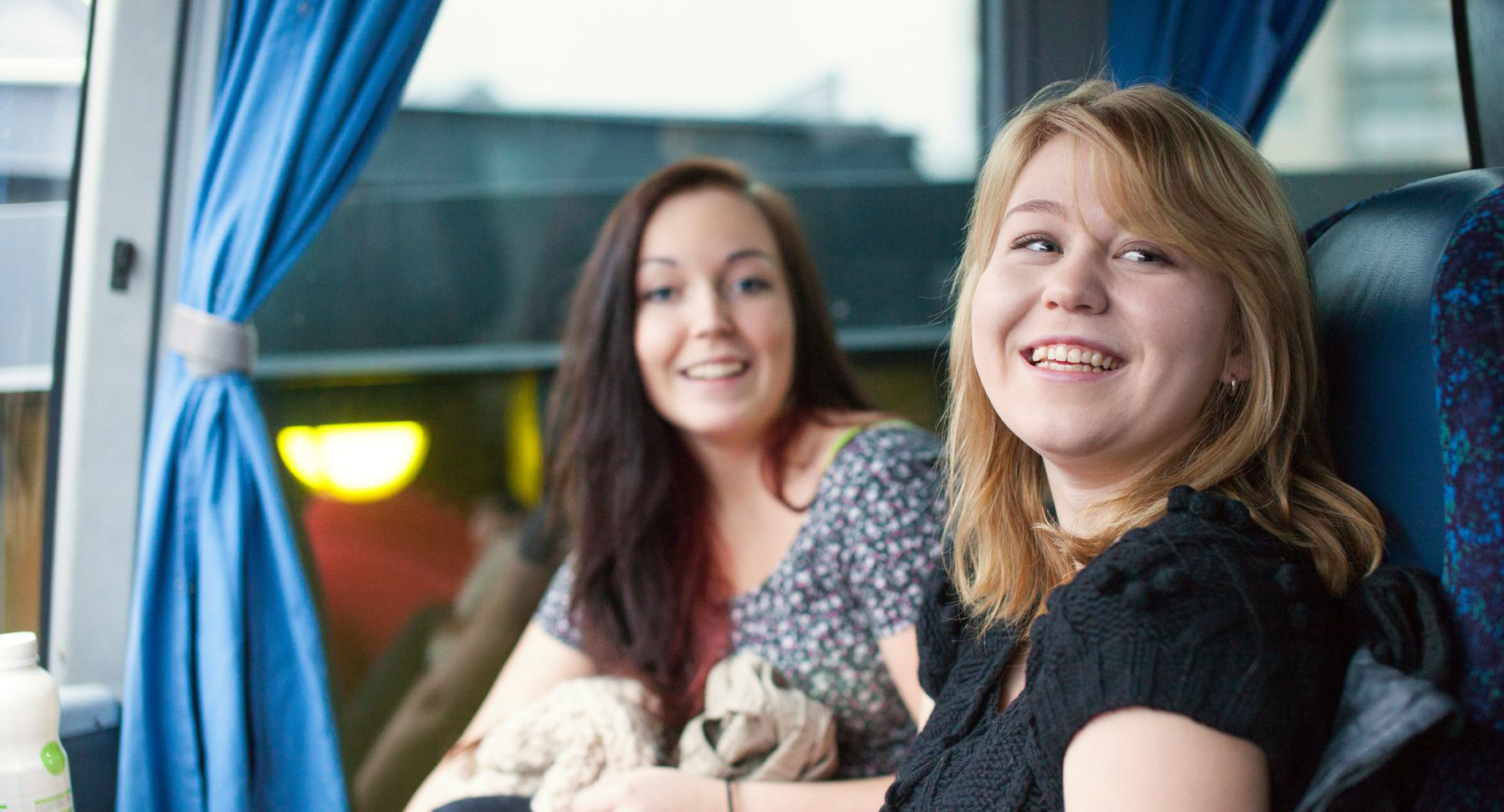 Två yngre kvinnor sitter tillsammans i en buss och skrattar mot kameran. 
