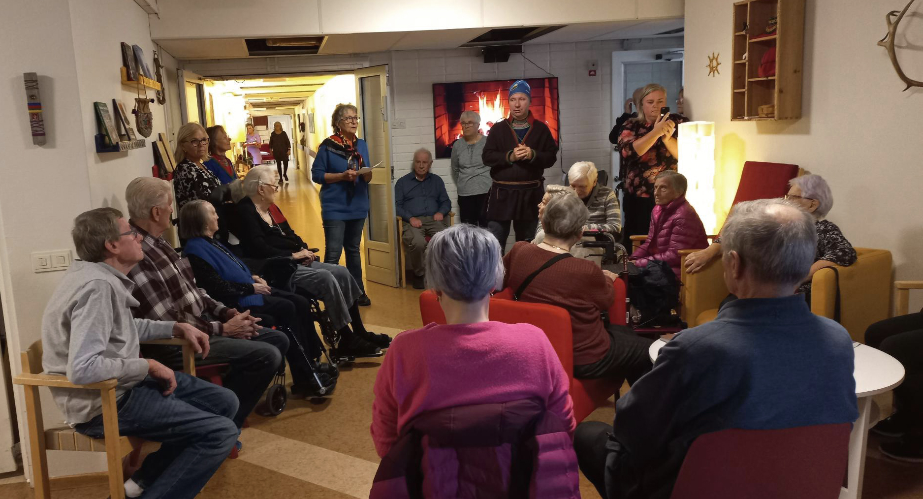 Bild på flera personer i grupp som lyssnar på en man som talar i samiska kläder.