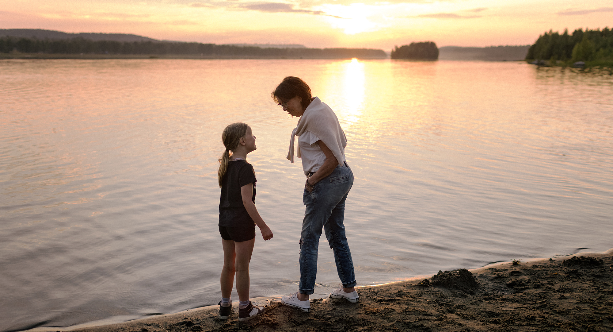Ett barn och kvinna står på en strand och skrattar och tittar på varandra i solnedgång.