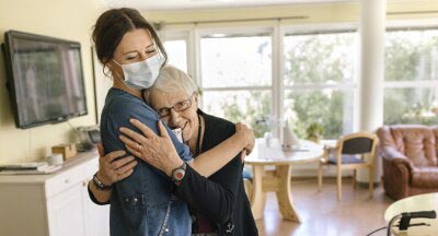 Bild på en kvinna med munskydd och vårdkläder som kramar en äldre kvinna. 