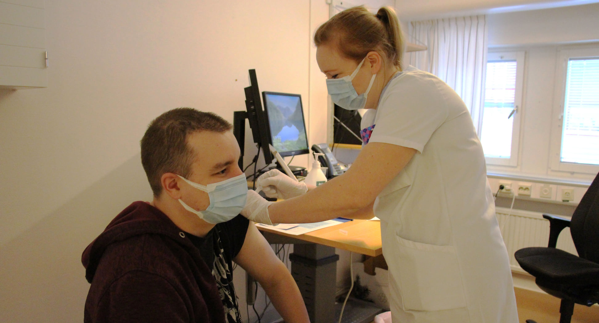 En kvinnlig sköterska i vita kläder tar en spruta på en yngre man men munskydd