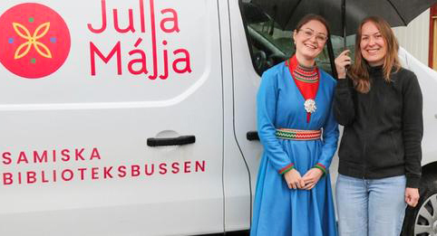 Två kvinnor står bredvid bussen Julla Majja och ler mot kameran. En i samisk dräkt. 