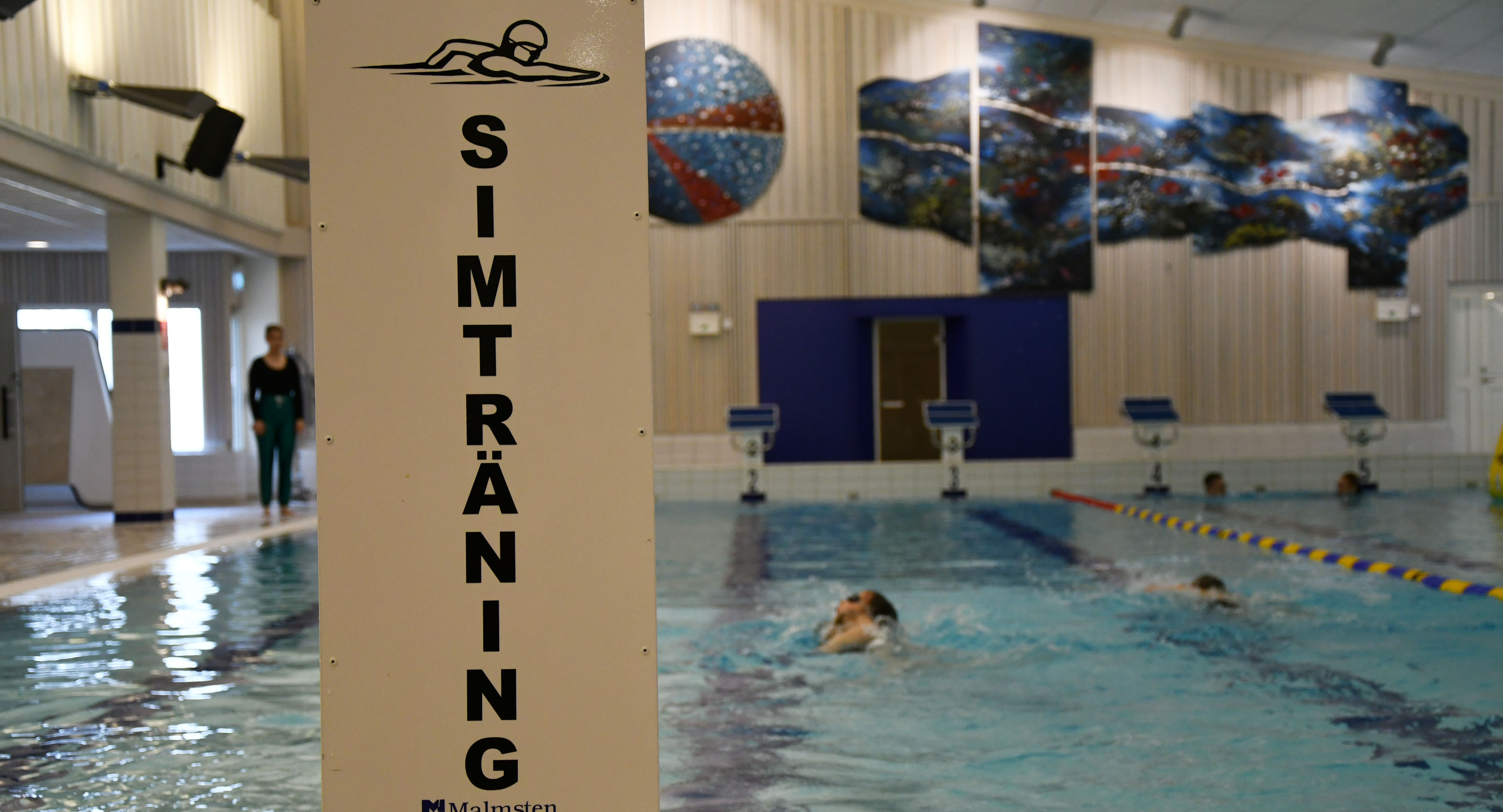 Skylt där det står simträning med person som simmar i bassäng